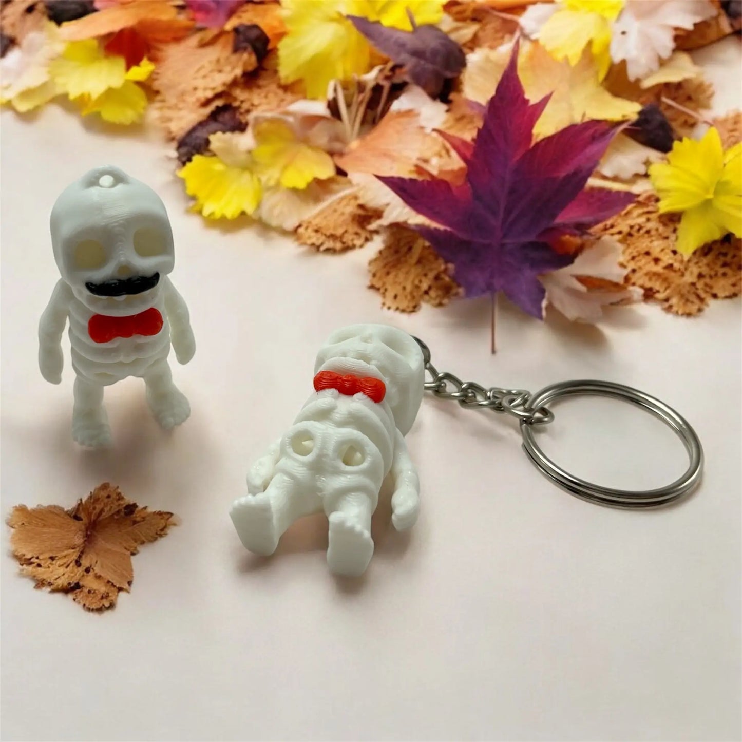Two white skeleton keychains with red bow ties on a white surface with autumn leaves.